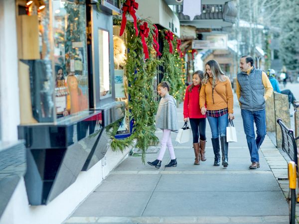 A family strolls through a festive shopping street, admiring a decorated Christmas garland and carrying holiday gift bags.