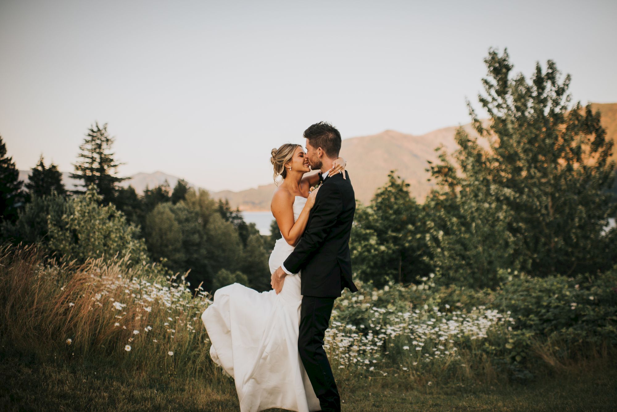 A couple embraces in wedding attire outdoors, surrounded by greenery and mountains in the background.