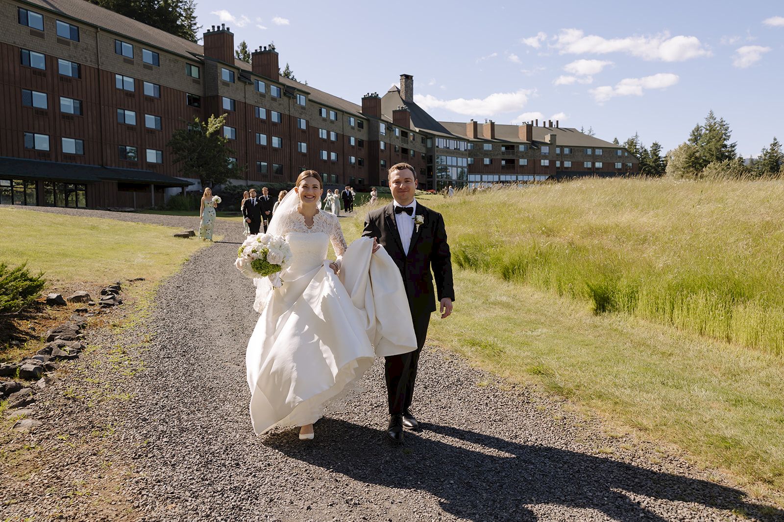 A bride and groom in formal wear walk along a gravel path outside a lodge on a sunny day.