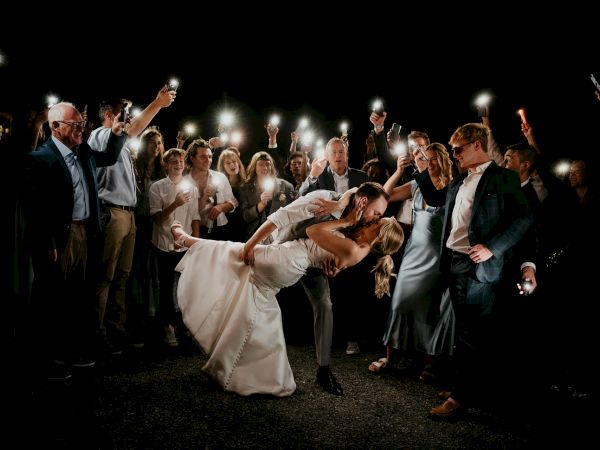 A couple kisses in the center while surrounded by a group holding up lights, creating a celebratory atmosphere.