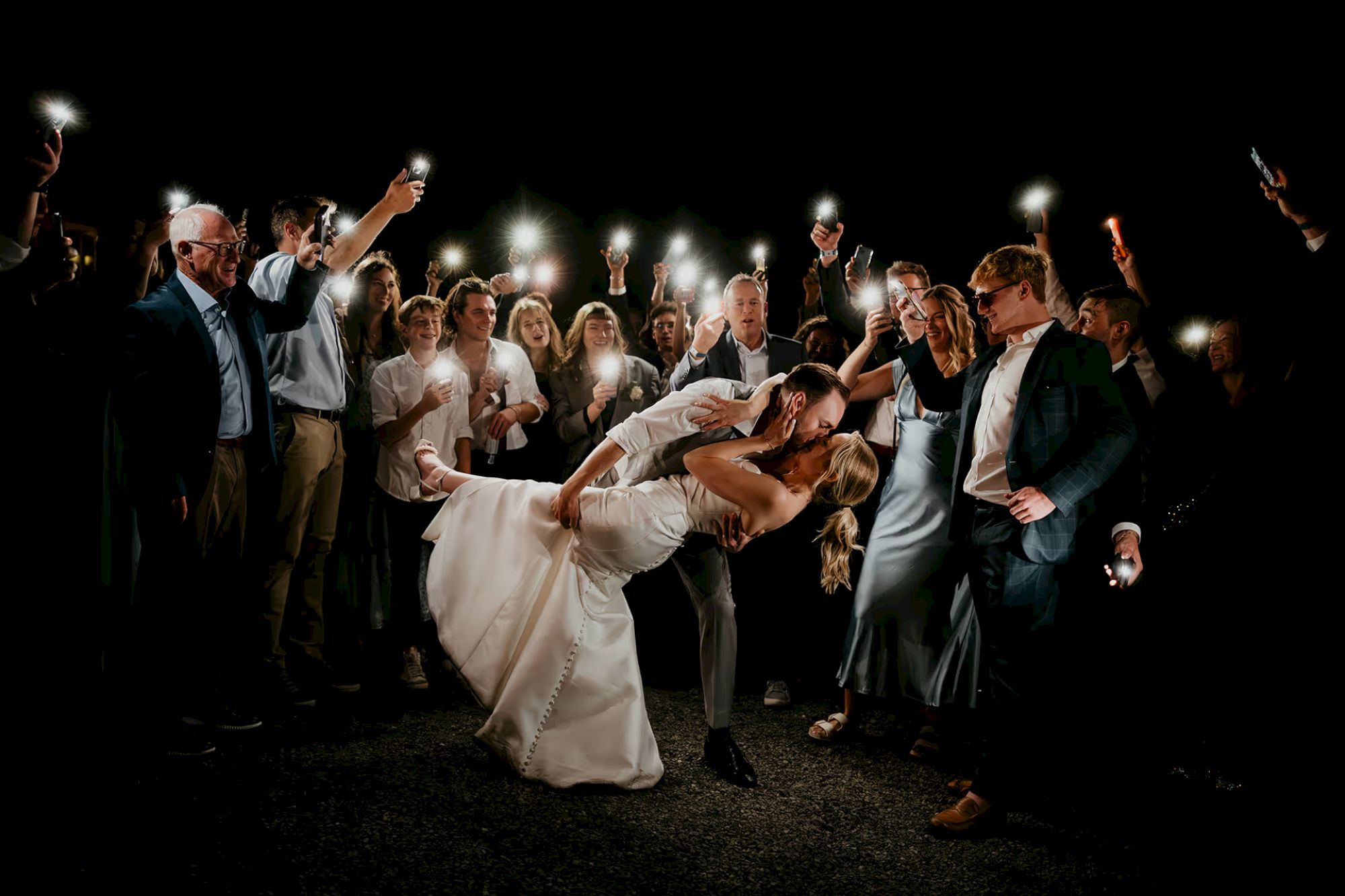 A couple kisses surrounded by a crowd holding up phone lights, creating a romantic nighttime scene.
