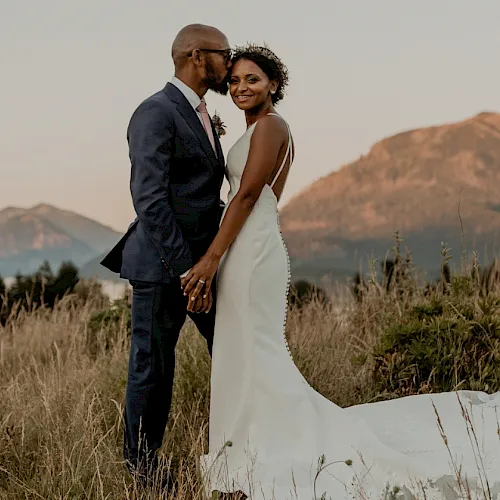 A bride and groom pose outdoors in a grassy field with mountains in the background, smiling and holding hands together at sunset.