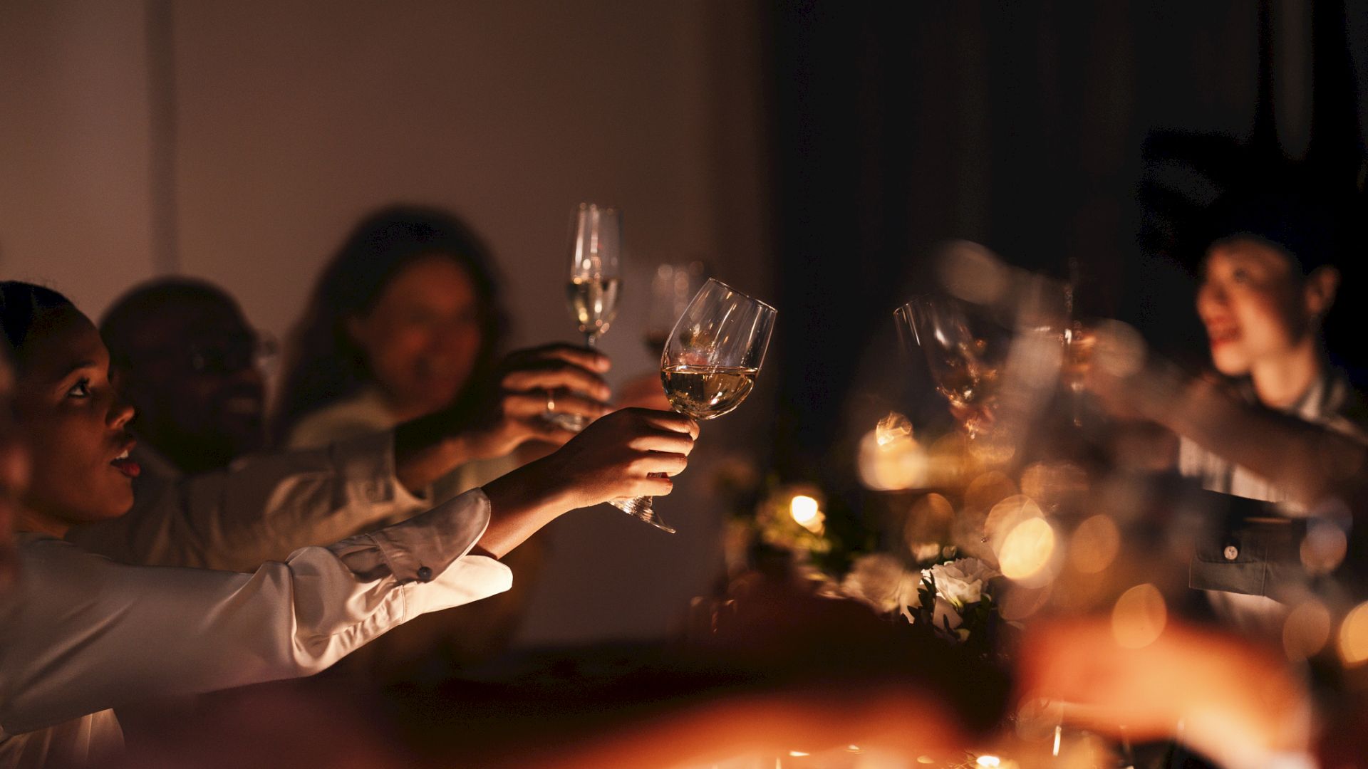 Friends clink champagne glasses at a candlelit celebration, toasting together in warm golden light.