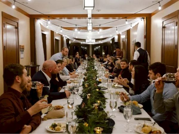 A long dining table with people enjoying a meal, set in a warmly lit room with decorative lights and a green centerpiece.