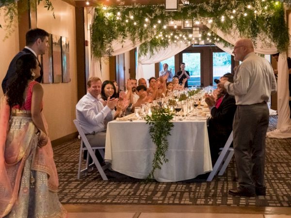 A formal gathering with people seated at a long table, one person standing and speaking, under string lights and greenery decorations.