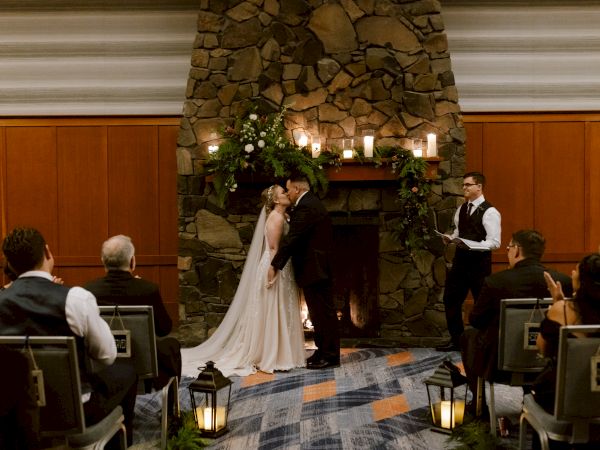 A couple is kissing at a wedding ceremony in front of a stone fireplace, with an officiant and guests present in an indoor setting.