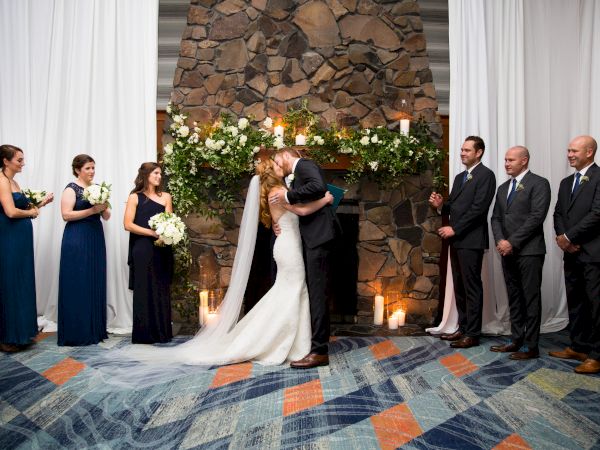 A bride and groom kiss at a wedding ceremony, surrounded by the bridal party. The backdrop is a stone wall with floral arrangements and candles.