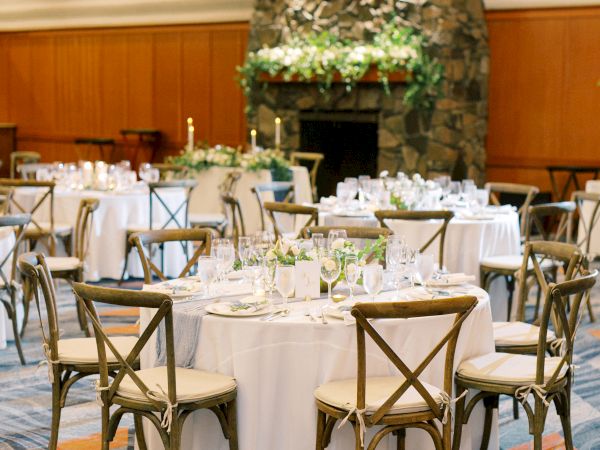 A decorated event space with round tables, white tablecloths, wooden chairs, and a stone fireplace adorned with greenery.