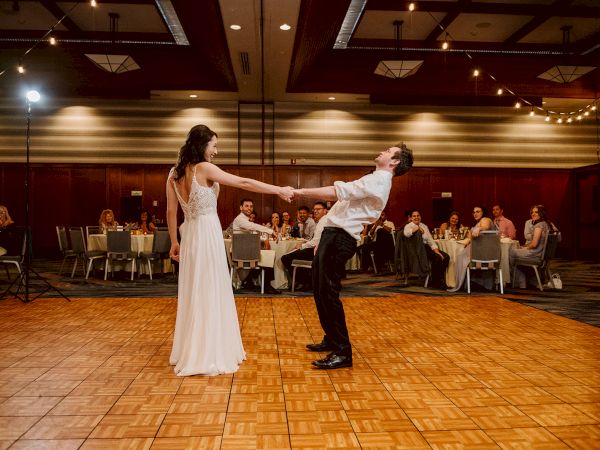 A couple dances energetically on a wooden floor in a decorated hall with seated guests watching and lights hanging above.