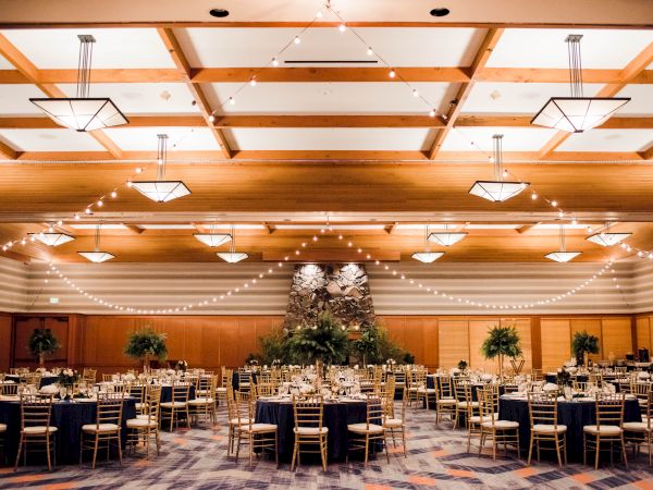 A decorated banquet hall with round tables, chairs, string lights, and a stone wall centerpiece, likely prepared for an event or reception.