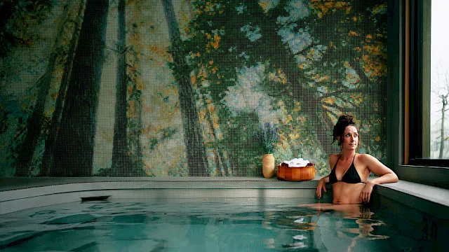 A woman relaxes in an indoor spa pool beneath a colorful mosaic wall, with towels and flowers on the ledge nearby.