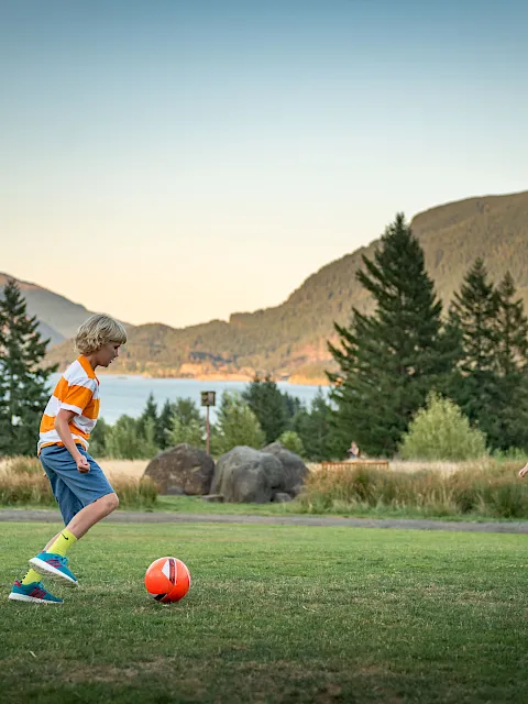 Kids play soccer on a grassy field by a scenic lake, with tall evergreens and mountains in the warm sunset backdrop.