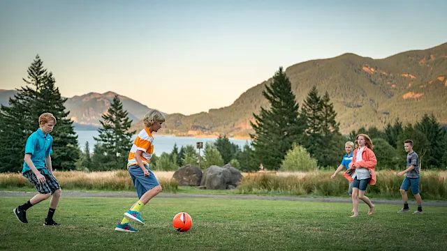 Kids play soccer on a grassy field by a scenic lake, with tall evergreens and mountains in the warm sunset backdrop.