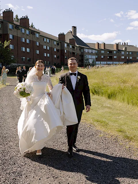 A bride and groom in wedding attire stroll down a gravel path beside a grassy lawn and a lodge building during daylight.