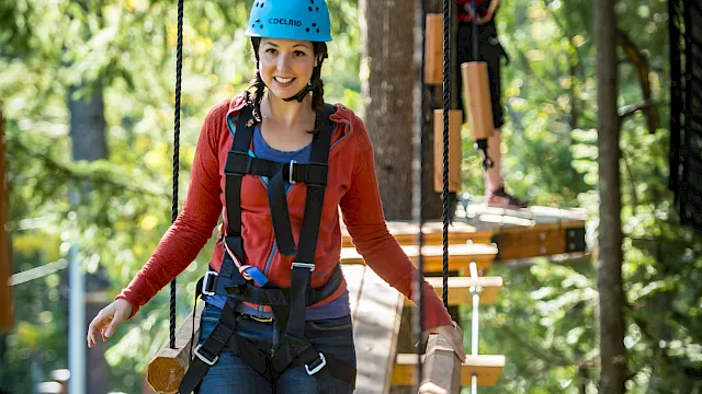 A woman in a blue helmet and safety harness stands on a rope bridge in a forest adventure park, smiling.