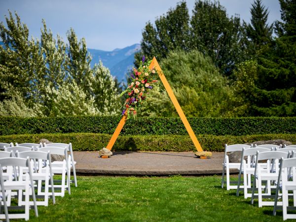An outdoor wedding setup with an empty aisle, white chairs, and a triangular wooden arch adorned with flowers and greenery.