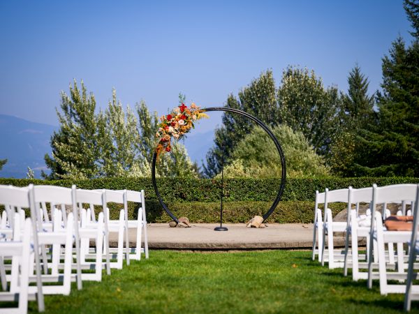 An outdoor wedding setup with white chairs facing a decorative circular arch adorned with flowers, set against a backdrop of trees and clear skies.