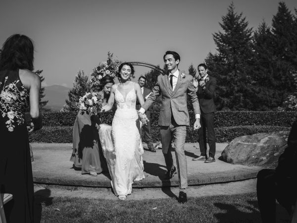 A newlywed couple walks down the aisle, smiling, outdoors with guests and a bridal party applauding.