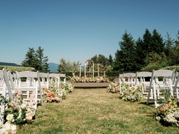 The image shows an outdoor wedding setup with white chairs, a floral aisle, and an arch at the front, surrounded by greenery and blue skies.