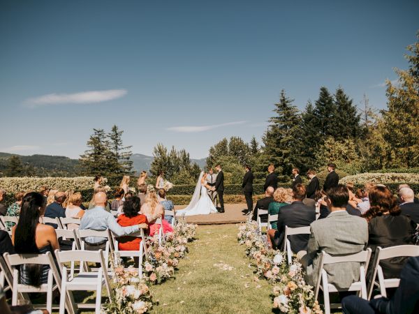 A wedding ceremony outdoors with guests seated and a couple at the altar surrounded by trees and greenery.