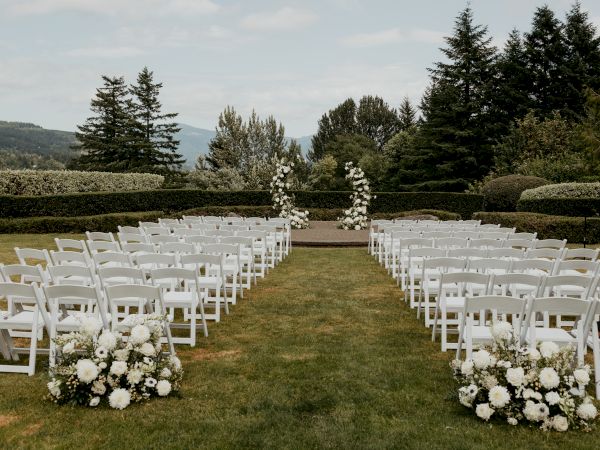 An outdoor wedding setup with white chairs and floral arrangements, set on a grassy area surrounded by trees and distant hills.