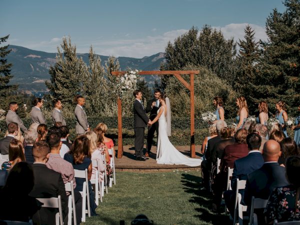 A couple is having an outdoor wedding ceremony with guests seated, surrounded by greenery and mountains in the background.