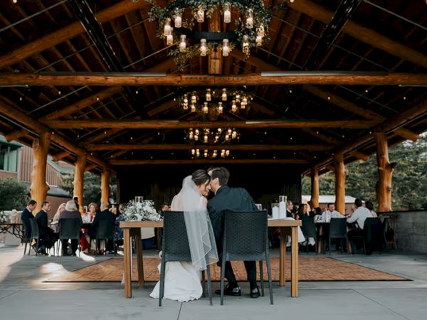 A couple in wedding attire sits at a table in a rustic venue with guests nearby, under a wooden ceiling with hanging lights.