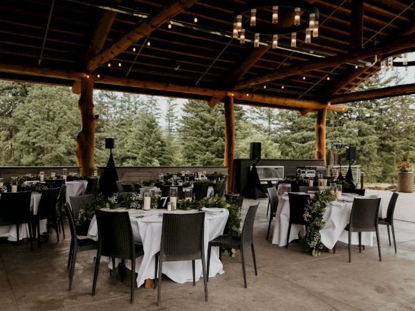 An outdoor event setup with round tables, white tablecloths, and black chairs under a wooden pavilion surrounded by trees in the background.