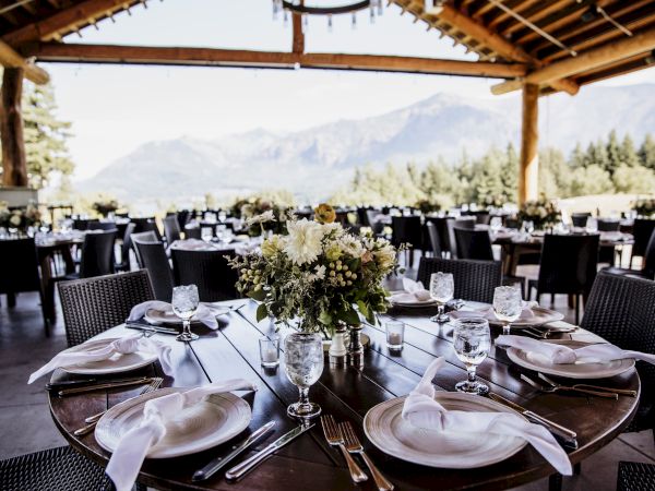 An elegant outdoor dining setup with floral centerpieces and mountains in the background, under a wooden roof.