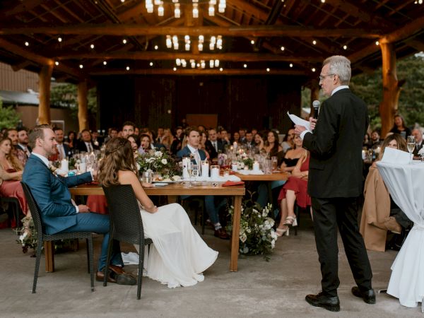 A wedding reception with guests seated at tables, a man giving a speech, and a couple at a decorated table under a wooden structure.