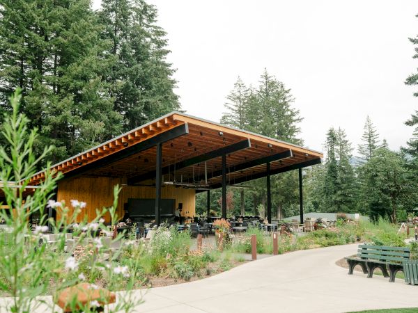 The image shows an outdoor pavilion with a wooden roof surrounded by trees, seating, and a garden-like setting.