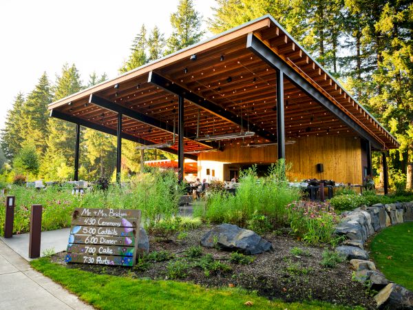 An outdoor venue with a wooden roof and string lights, surrounded by greenery. A schedule on a sign lists events like dinner and live music.