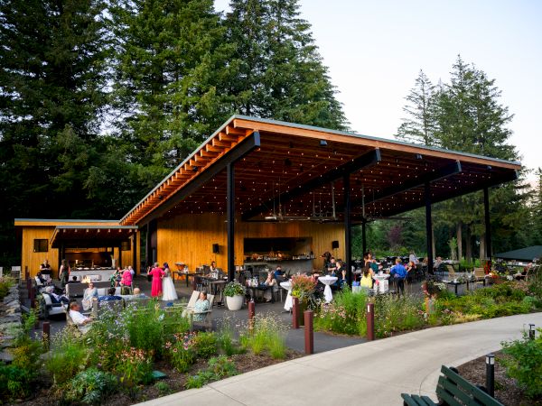 An outdoor restaurant with a wooden roof, surrounded by greenery and people dining and socializing.