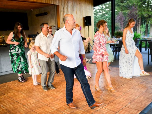 A group of people dancing indoors on a wooden floor, dressed in summer outfits, with a bar and outdoor view in the background.