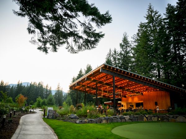 A pavilion with warm lighting in a wooded area, surrounded by trees and greenery, with a path leading towards it at dusk.