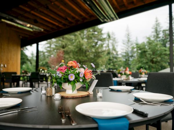 An outdoor dining setup with a table adorned with a floral arrangement, plates, and cutlery, surrounded by trees under a wooden roof.
