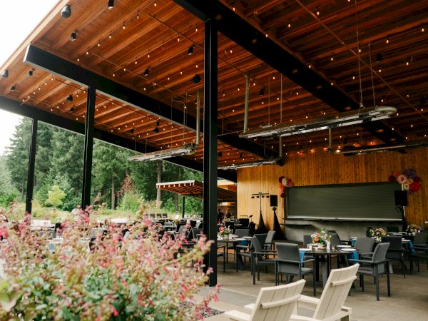 An outdoor restaurant patio with wooden ceiling, string lights, and empty tables and chairs surrounded by greenery and plants.