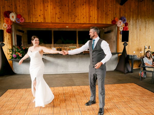 A couple is dancing on a wooden floor. The woman wears a white dress, and the man is in a suit. The setting is a decorated hall.