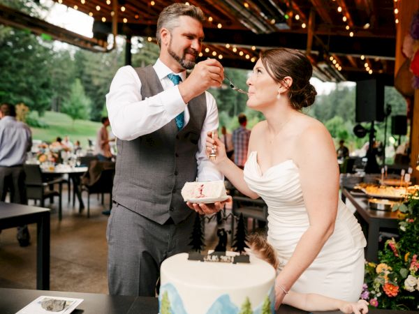 A couple is at a wedding reception, and the groom is feeding cake to the bride, surrounded by guests and decorated tables.