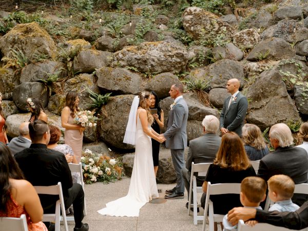 A wedding ceremony is taking place outdoors, with a couple exchanging vows in front of guests seated on white chairs.