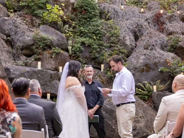 A wedding ceremony is taking place outdoors with guests seated, a couple, and an officiant.