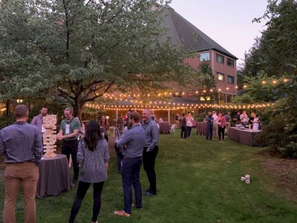 A group of people are enjoying an outdoor gathering, playing giant Jenga under string lights on a grassy area.