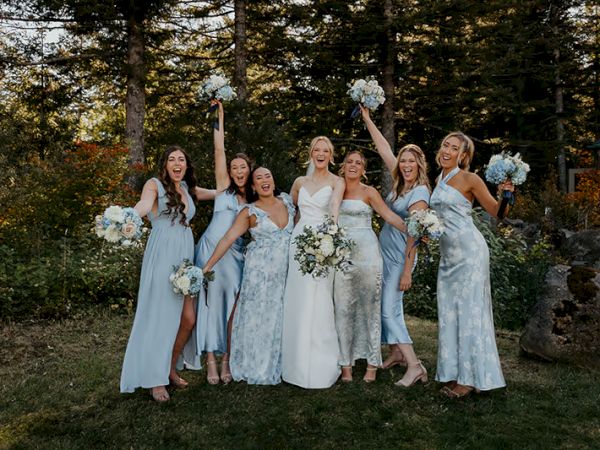 A group of women in dresses pose joyfully outdoors, holding bouquets, surrounded by trees and greenery, celebrating an event.