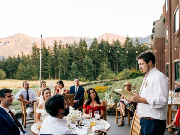 A group of people sit outside at wooden tables, listening to a man giving a speech. Trees and mountains are in the background.