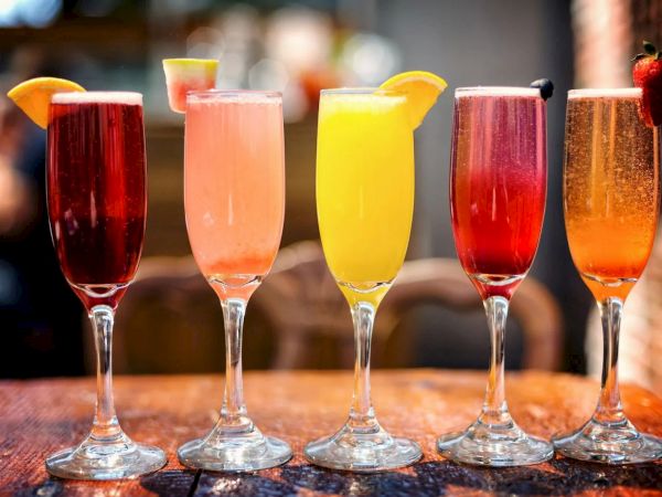 Five colorful cocktails in fluted glasses, each with different garnishes like fruits, arranged on a wooden table.