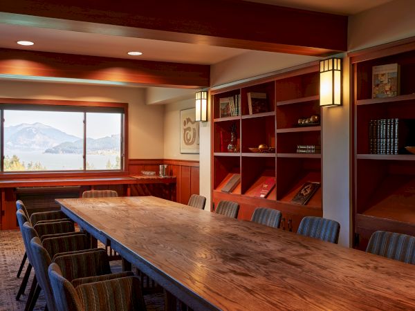 A conference room with a wooden table, chairs, bookshelves, and a window revealing a mountain view under warm lighting.