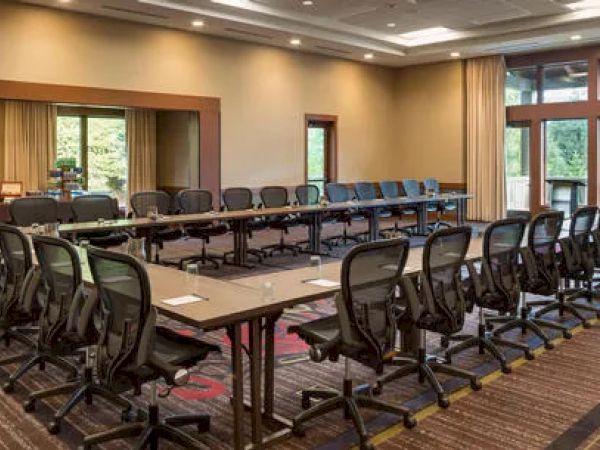 A large conference room with U-shaped tables, office chairs, and a beverage station, surrounded by windows with natural light.