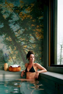 A person sits in a tranquil indoor pool, relaxing by the edge with a drink, a peaceful forest mural on the wall behind, and large windows letting in light.