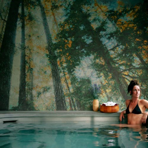 A person sits in a tranquil indoor pool, relaxing by the edge with a drink, a peaceful forest mural on the wall behind, and large windows letting in light.