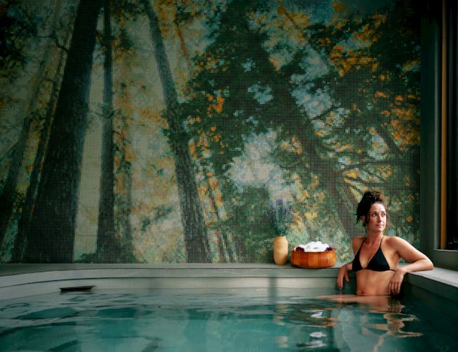 A person sits in a tranquil indoor pool, relaxing by the edge with a drink, a peaceful forest mural on the wall behind, and large windows letting in light.